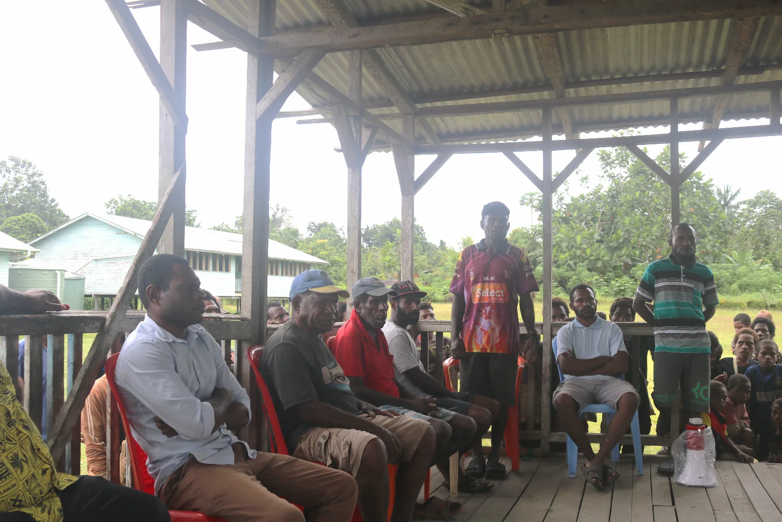 Community members taking part in a meeting in an open-sided shelter.