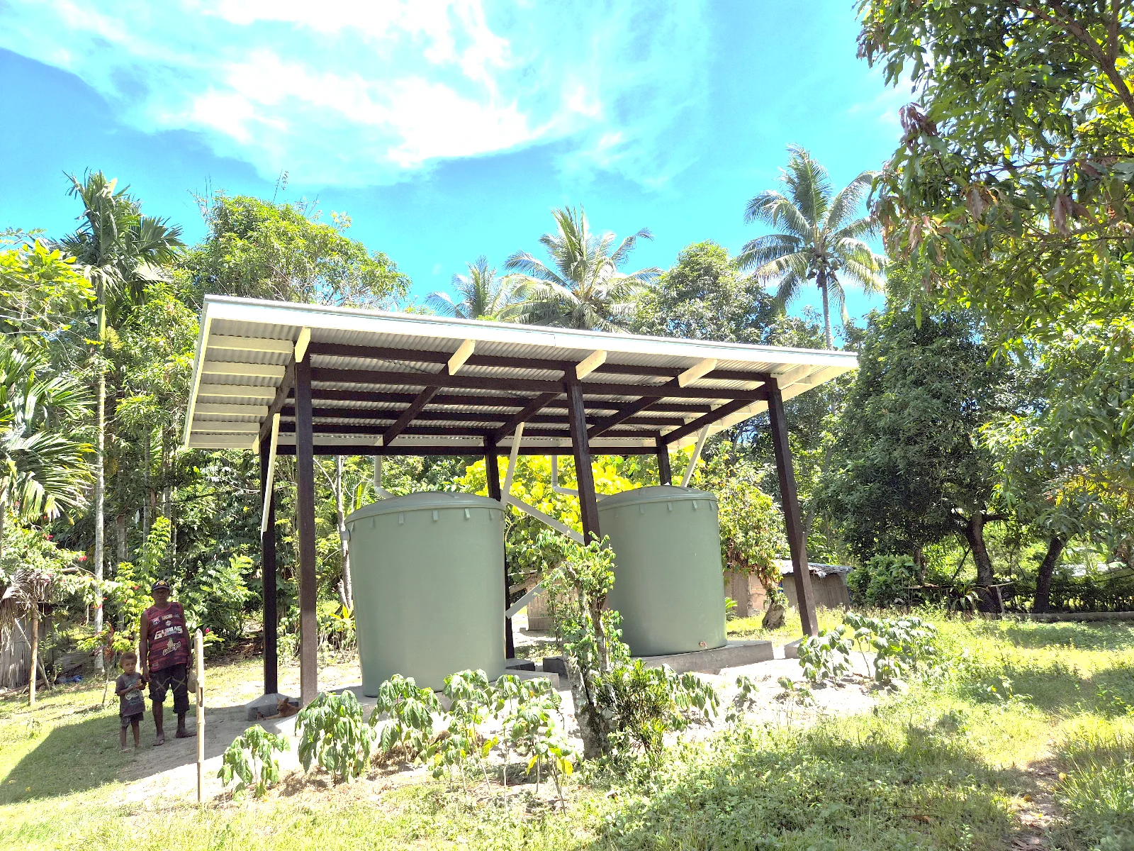 Rainwater tanks beneath a roofed catchment structure, surrounded by tropical vegetation.